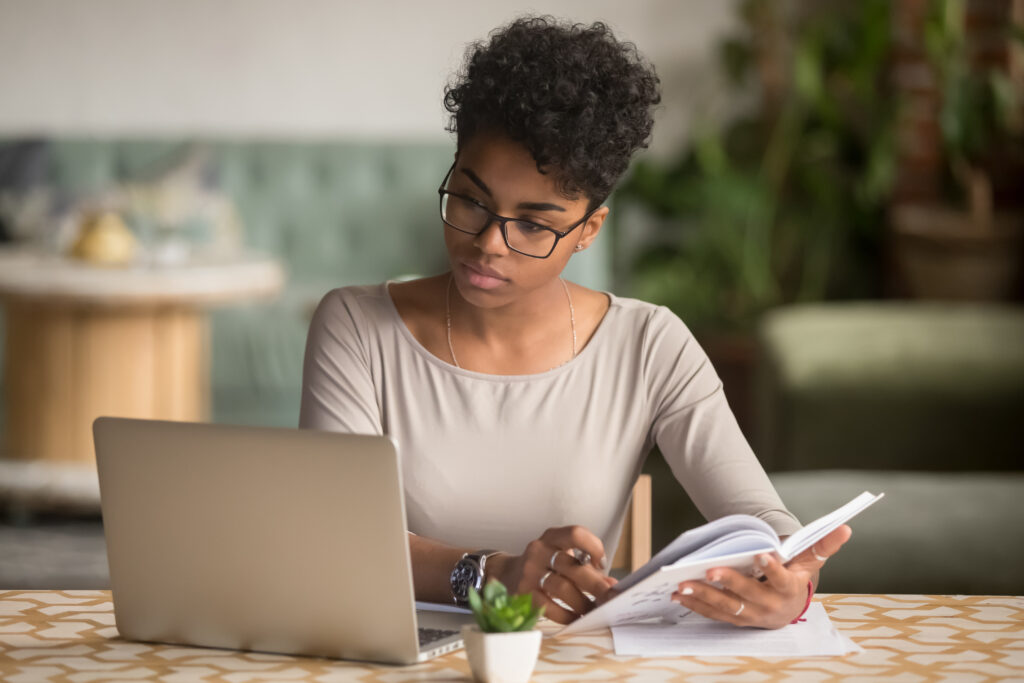 Black woman at a table looking at a computer and holding a book to see if she is ready to start an ERG program