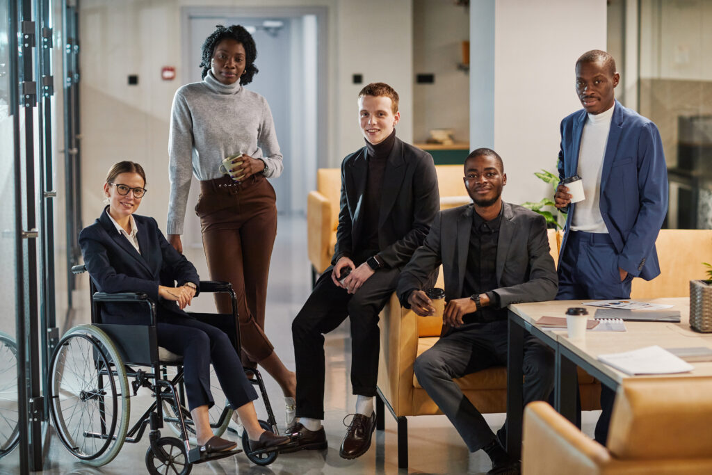 Full length portrait of diverse business team with young woman in wheelchair all smiling at camera in office