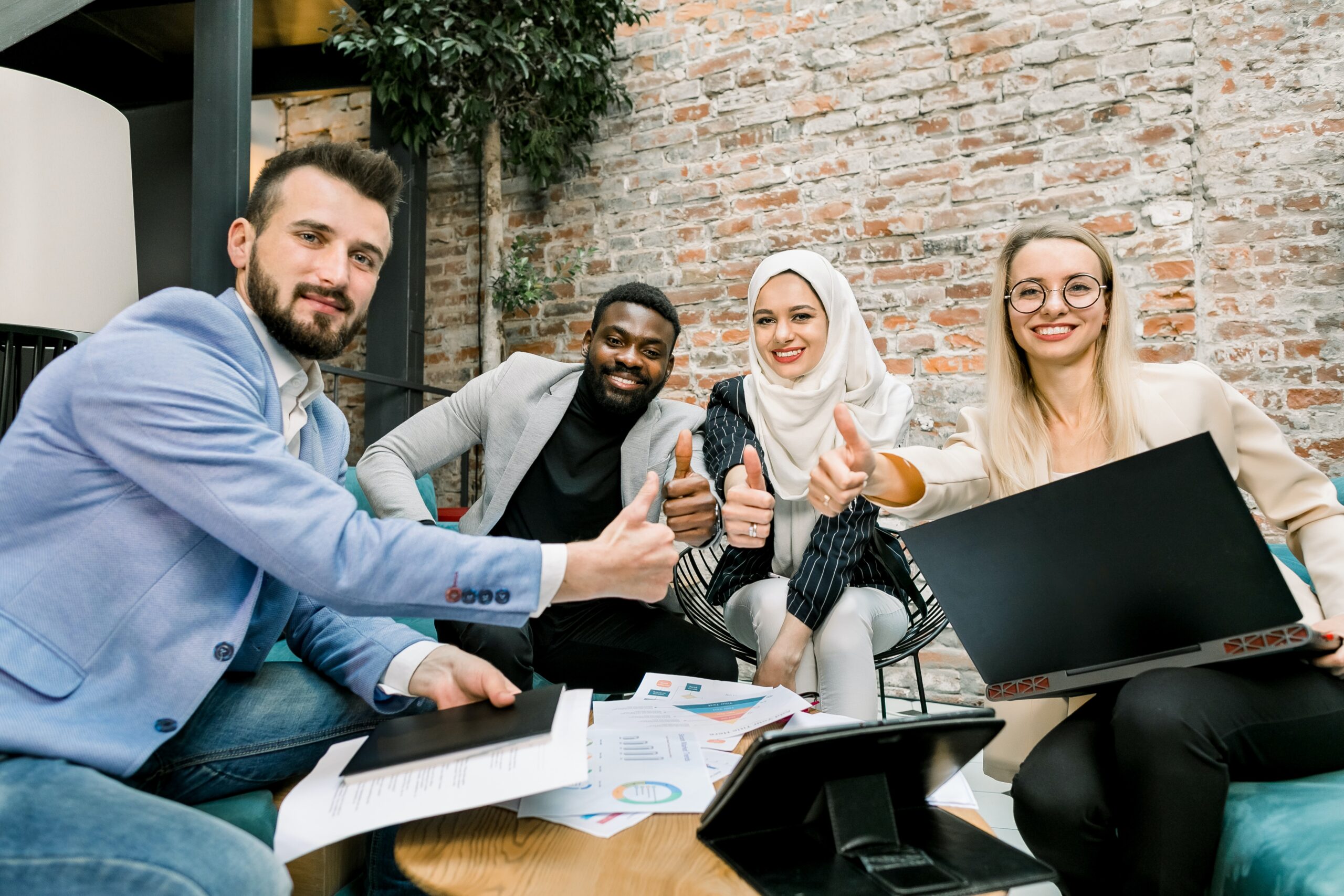 A diverse group of four people sitting around a table with papers on them smiling and giving a thumbs up