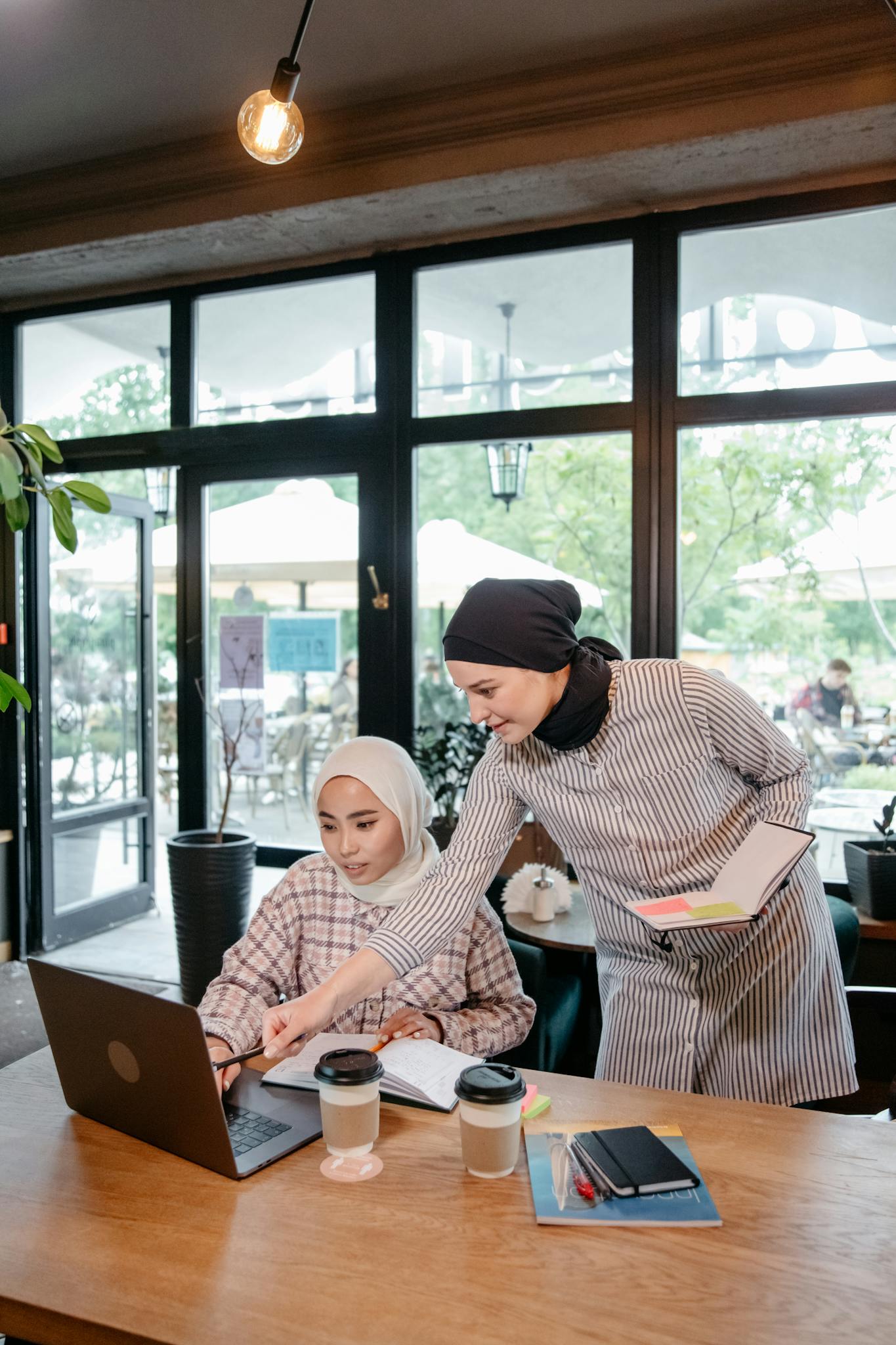 Two Muslim women collaborate over a laptop in a cozy café, discussing business ideas.